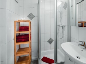 Une salle de bain avec une étagère en bois où sont posées des serviettes rouges, une douche en verre et un lavabo blanc.