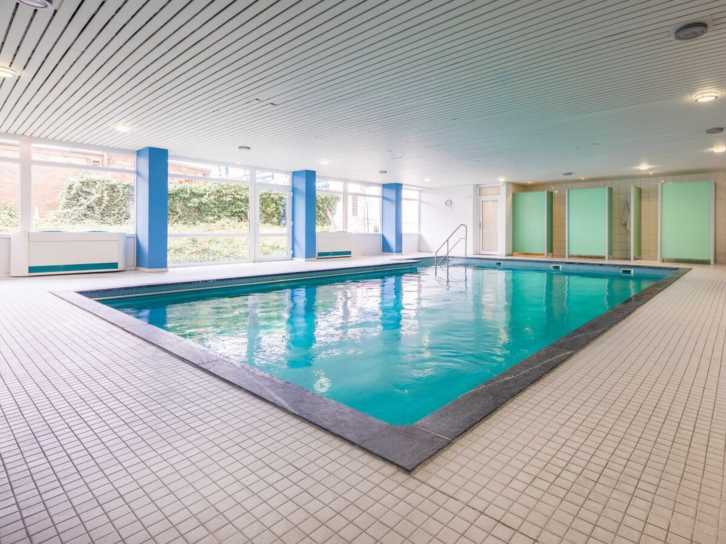Ein beheiztes Hallenbad mit Fliesenboden, blauer Beckenumrandung und Deckenbalken in Weiß.