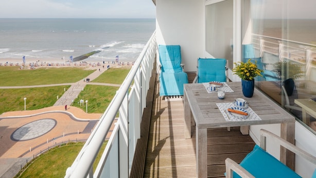 A balcony with a view of the beach, a table with a potted plant, and blue cushioned seating.