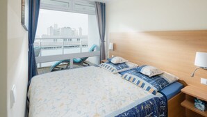 A hotel room with a bed, a nightstand, a lamp, a chair, and a view of the city.
