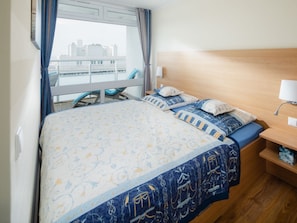 Habitación de hotel con cama, mesita de noche, lámpara, silla y vista a la ciudad.