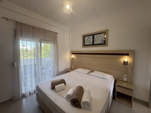 A hotel room with a bed, a nightstand, a wall-mounted light, a window with curtains, and a view of greenery outside. - Makis House (Sithonia)