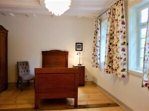 Ein Schlafzimmer mit einem hölzernen Bett, einem Sessel, einem Nachttisch mit Lampe und einem Fenster mit floralem Vorhang.