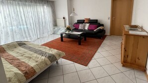 A bedroom with a bed, a coffee table, a sofa, a wooden cabinet, and a window with curtains. - Waibel Apartment, 60 sqm, 1 Bedroom, Terrace, Sleeps up to 3 Guests (Überlingen)