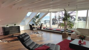 A modern living room with a black leather chair, a sofa, a dining area with a table and chairs, and a large window with a view of the outside. - Apartment 'Bodenseeblick' with Lake View, Balcony and Wi-Fi (Nonnenhorn)