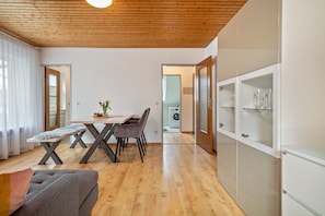 A modern living room with a wooden ceiling, a dining table with chairs, a sofa, and a cabinet with glass doors.