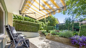 Un espace patio doté d’un coin salon abrité, d’un bac à fleurs en béton et d’une haie.