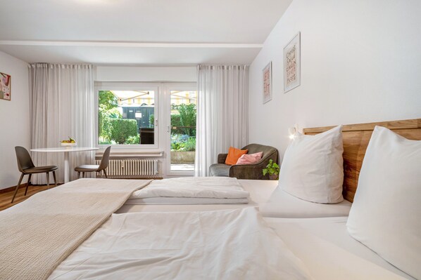 A hotel room with a bed, a desk, two chairs, a window with curtains, and a view of greenery outside.