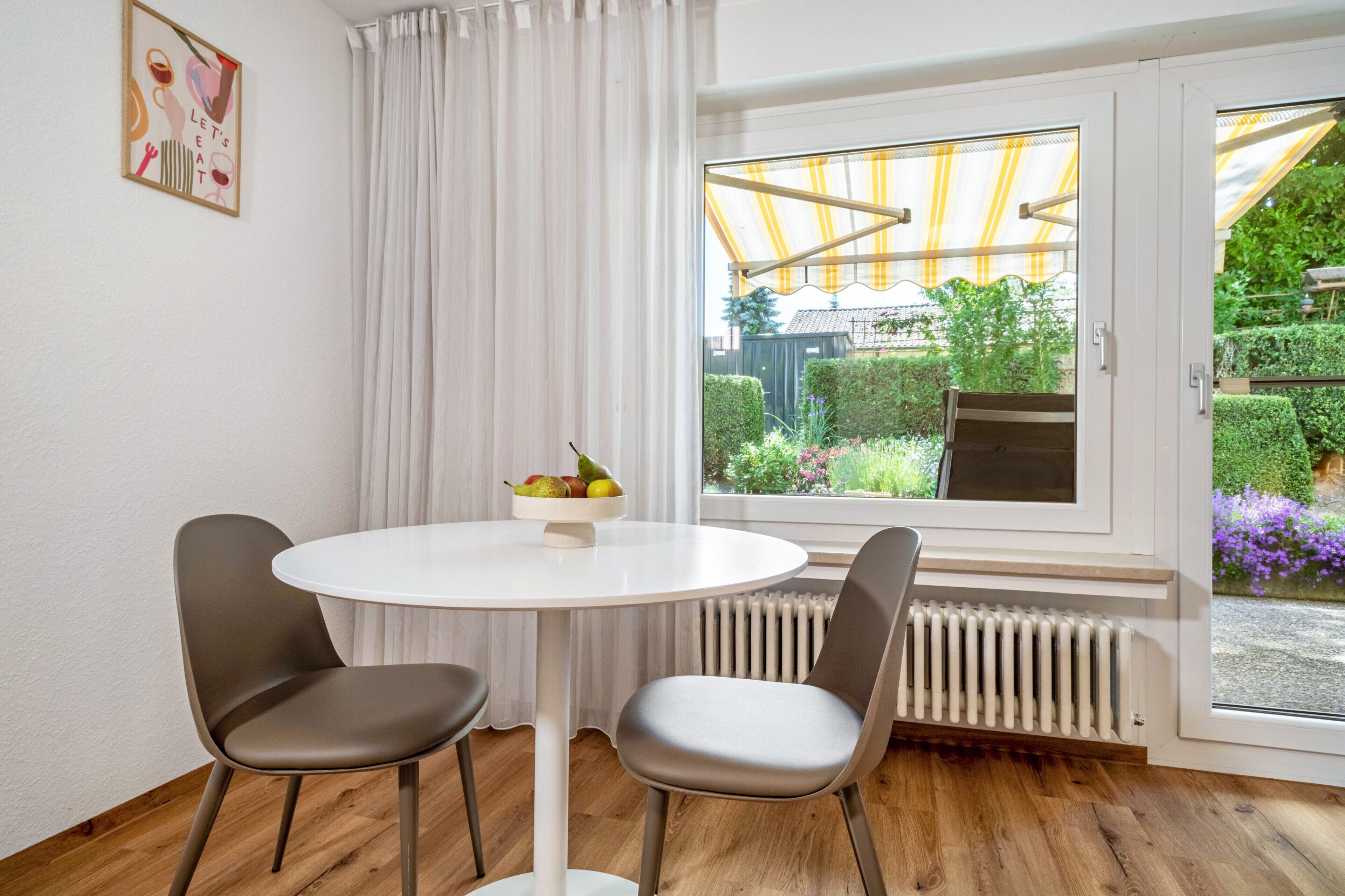 Ein Zimmer mit einem runden weißen Tisch, zwei grauen Stühlen, einem Fenster mit Blick auf Grünflächen, einem Heizkörper und einem Vorhang.