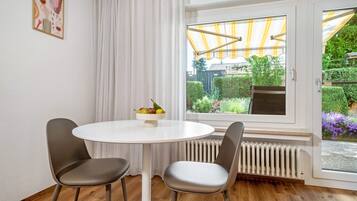 Ein Zimmer mit einem runden weißen Tisch, zwei grauen Stühlen, einem Fenster mit Blick auf Grünflächen, einem Heizkörper und einem Vorhang.