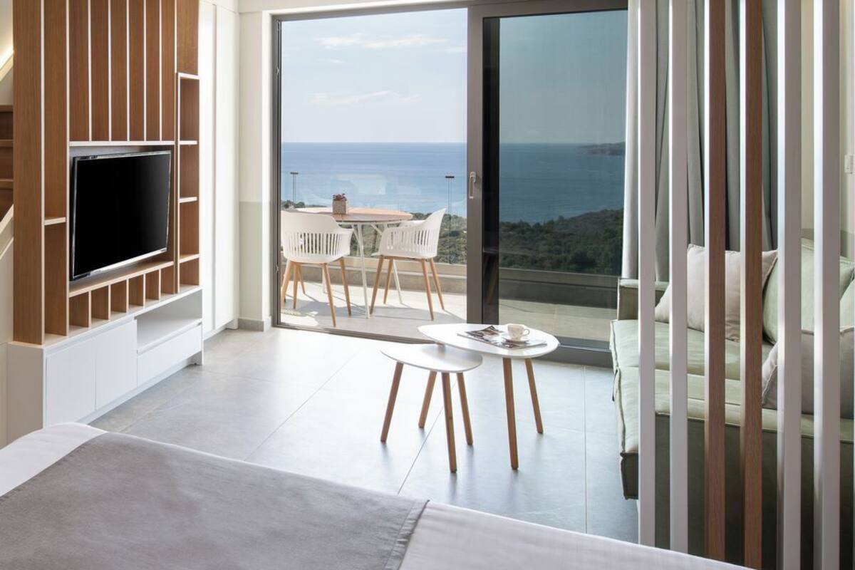 A modern living room with a flat-screen TV, a dining area with a table and chairs, and a large sliding glass door leading to a balcony with a sea view.