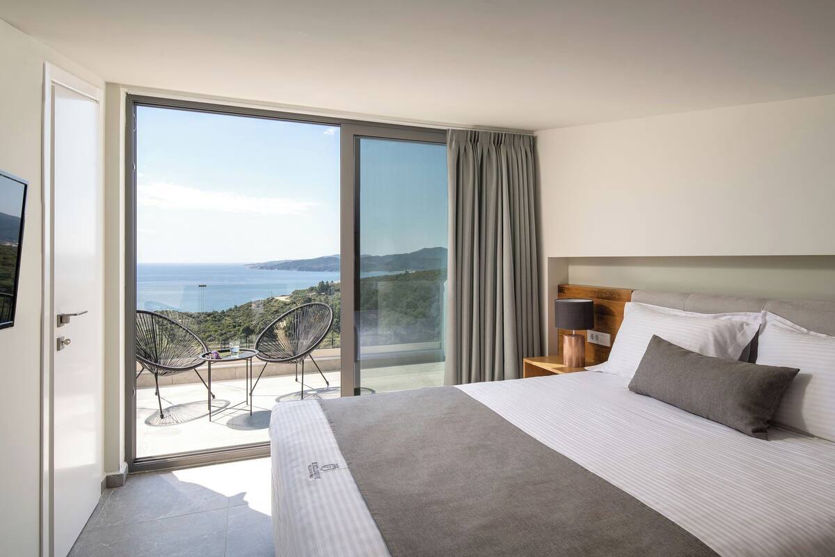 A hotel room with a large bed, a nightstand, a lamp, and a view of the sea through a sliding glass door.
