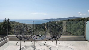 Un balcón con dos sillas y una mesita, con vistas a un paisaje costero con colinas y el mar.