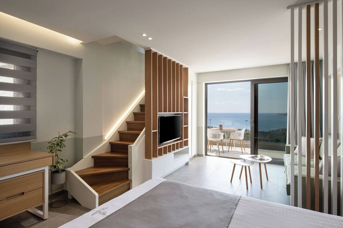 A modern living room with a wooden staircase, a flat-screen TV, a dining area with white chairs, and a large glass door leading to a balcony with a view of the sea.