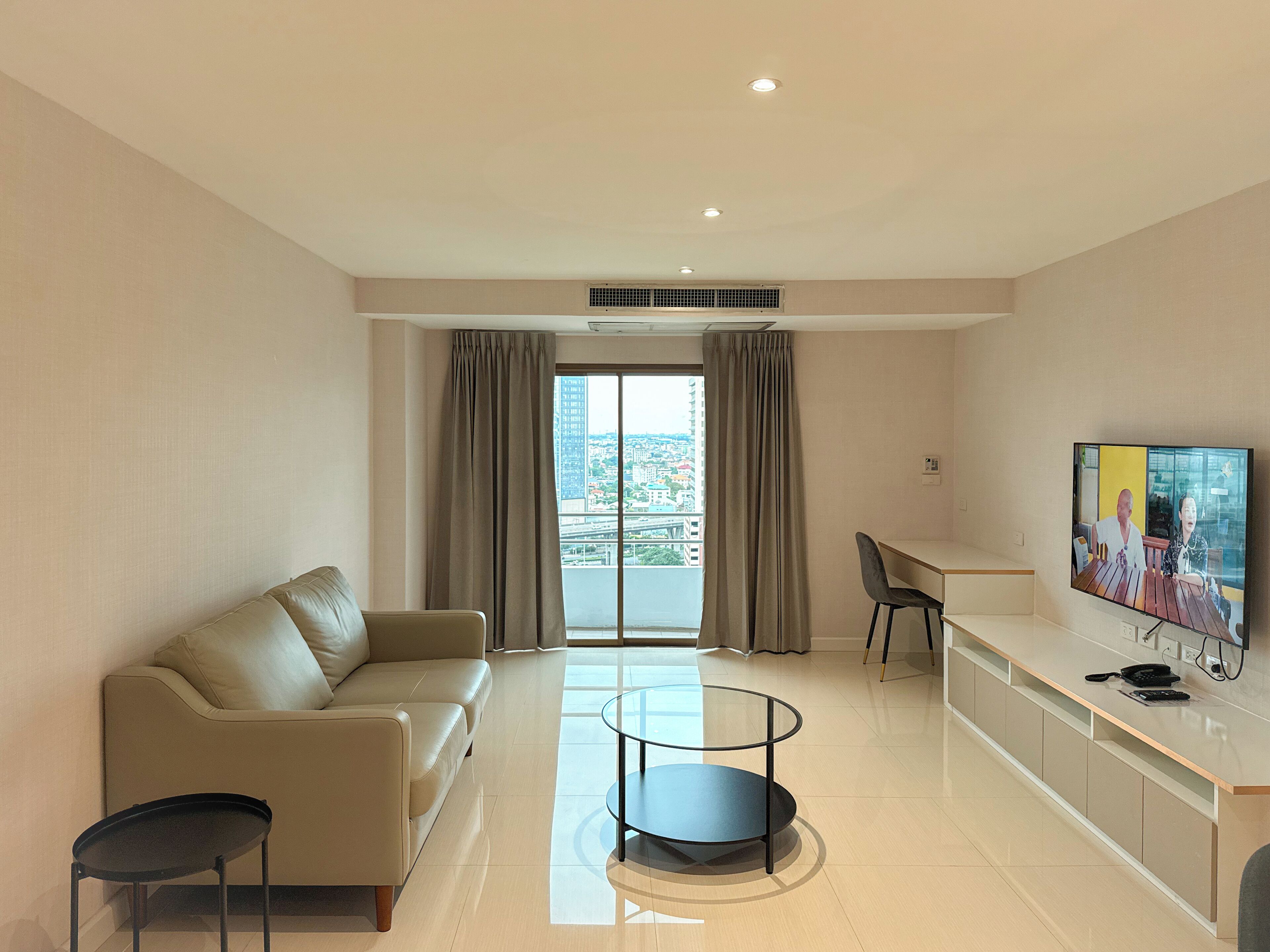 A modern living room with a sofa, a round coffee table, a TV, and a balcony with a city view.