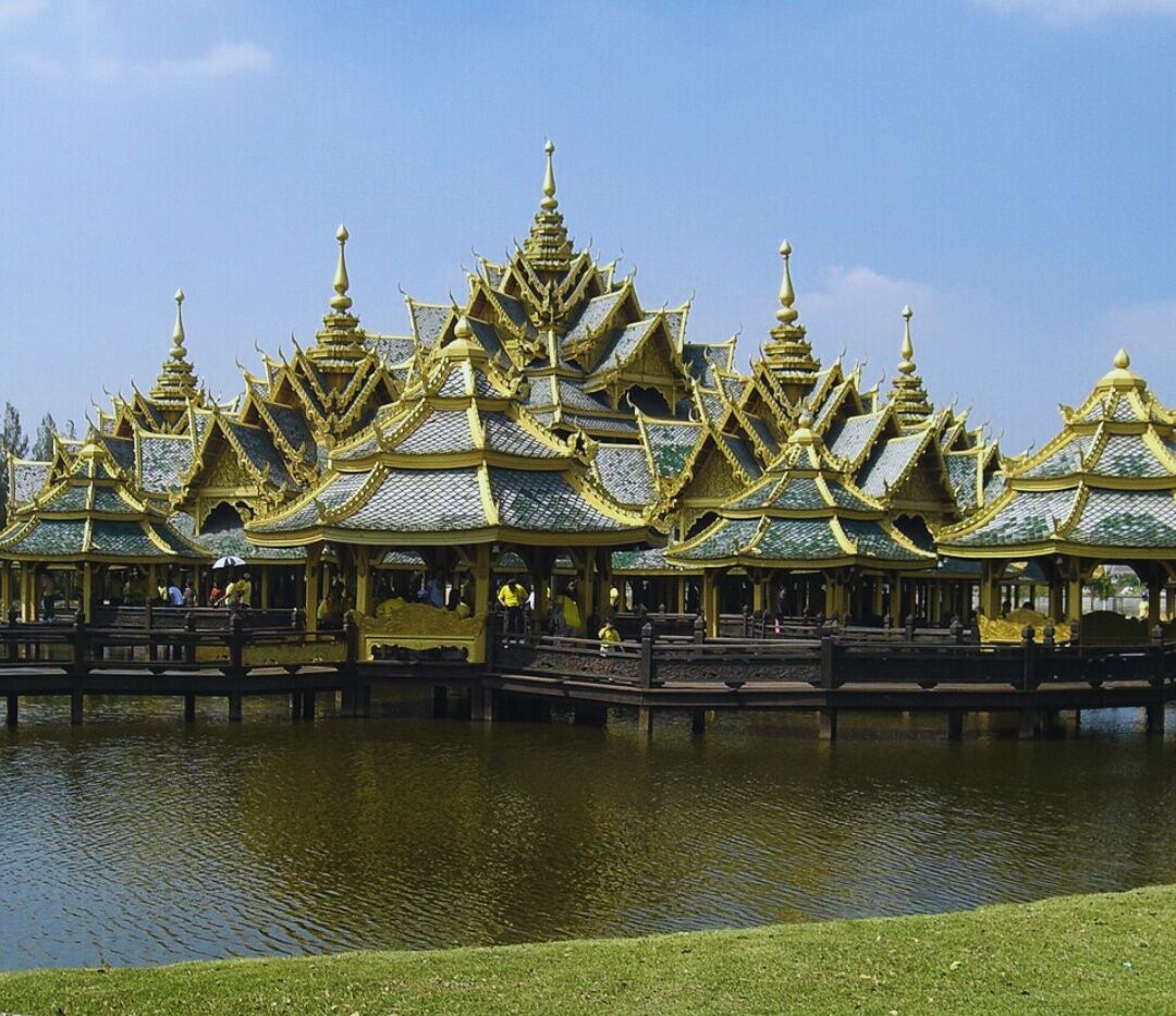 Une série de bâtiments de style thaïlandais traditionnel, dotés de toits richement décorés et surplombant un bassin d’eau aux reflets chatoyants.