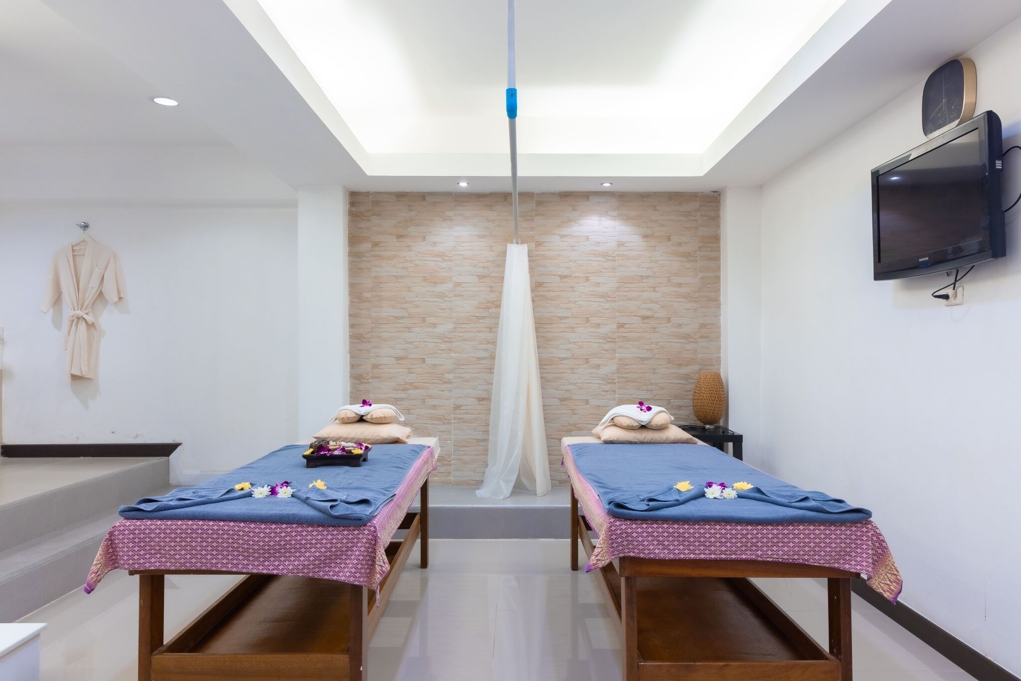 A modern spa room with two massage tables, a ceiling light, a TV, and a stone wall.