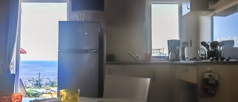 A compact kitchen with a refrigerator, a coffee maker, and a view of the sea through a window.