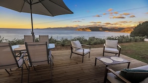 Una terraza de madera con mobiliario exterior, incluyendo sillas y una mesa, con vistas a un cuerpo de agua y montañas al fondo.