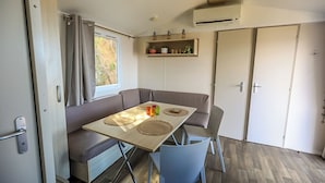 Un espacio habitable compacto con un sofá, una mesa con individuales, una silla azul, una ventana con cortinas, un estante con adornos y una puerta.