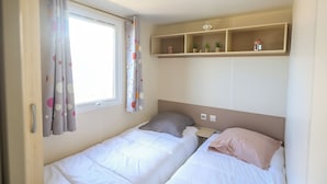Un alojamiento pequeño y de una sola habitación con dos camas, una ventana con cortinas, un estante con objetos pequeños y una lámpara en el techo.