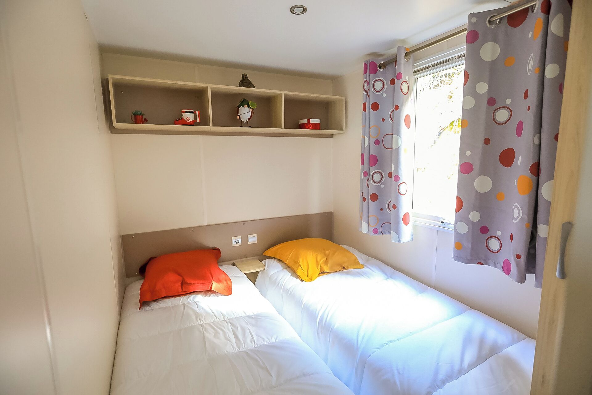 Un lit simple avec une literie blanche et deux oreillers, un orange et un jaune, dans une pièce dotée d’une fenêtre recouverte de rideaux à pois.