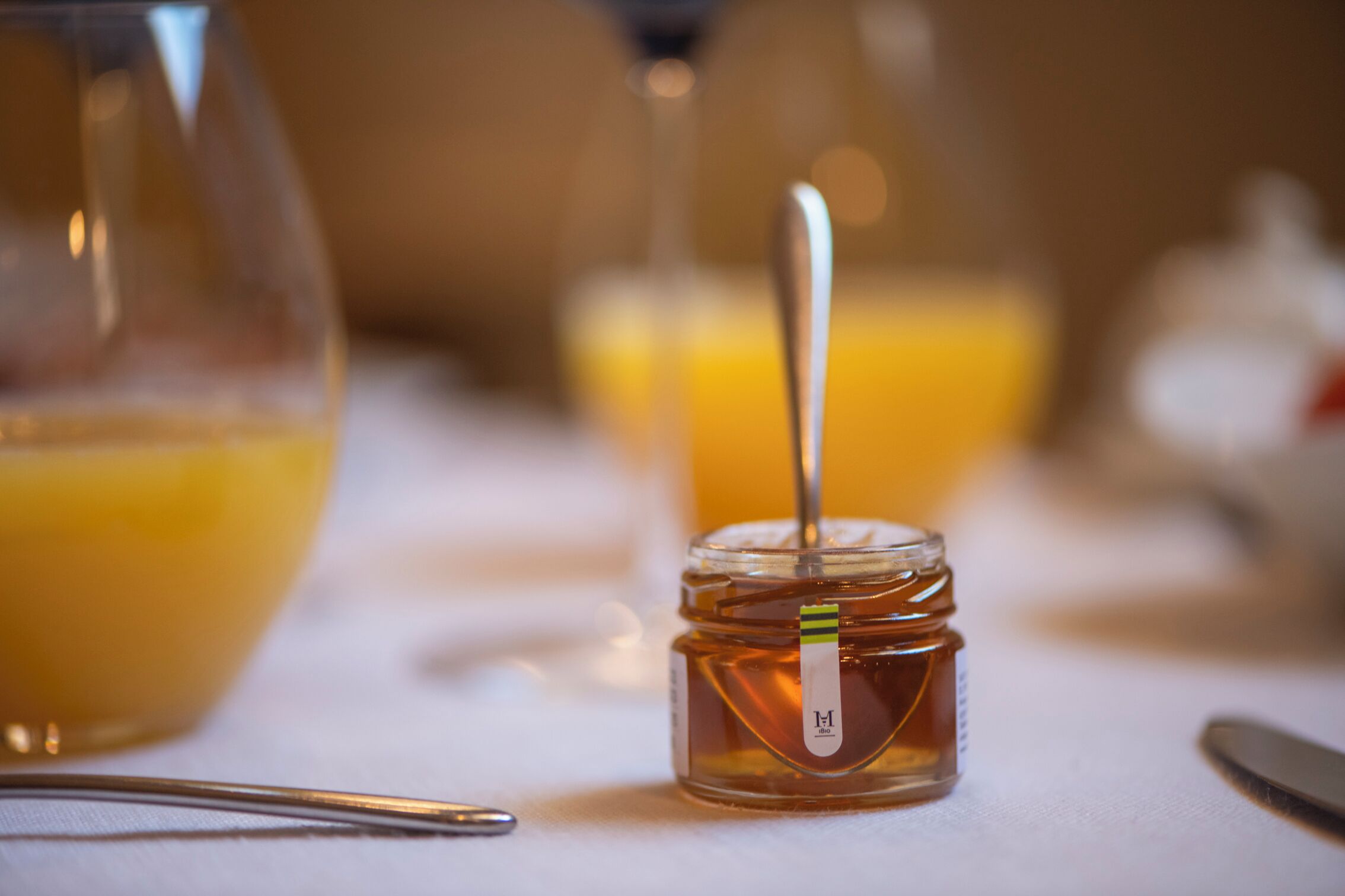 Primer plano de un tarro de miel con una cuchara, sobre una mesa junto a un vaso de jugo de naranja y un cuchillo.