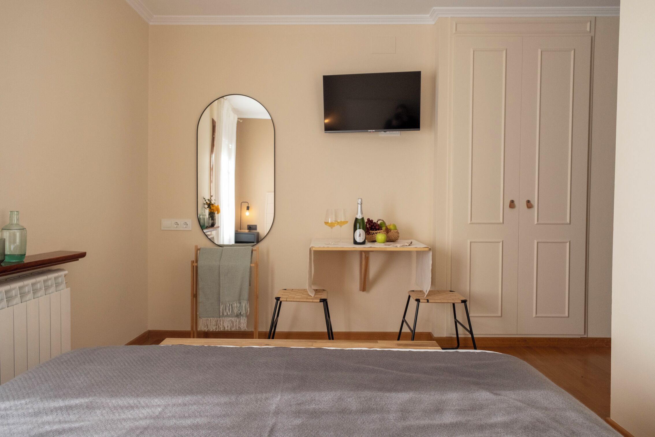 Une chambre avec un lit, un téléviseur fixé au mur, un miroir, une petite table avec une bouteille et des fruits, et une porte.