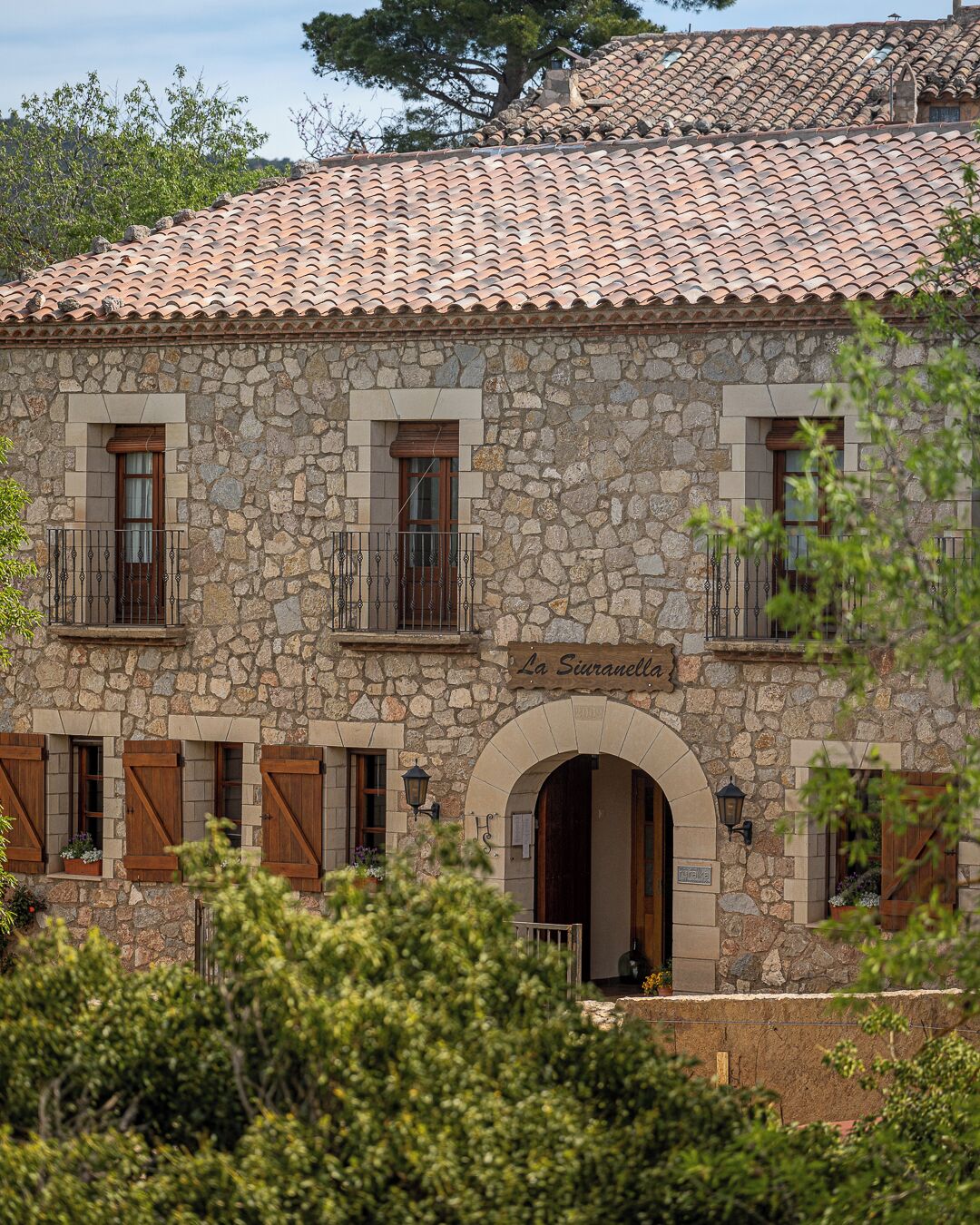 A stone building with a tiled roof, named "La Siuranella." It features multiple windows with wooden shutters, a central arched entrance, and a balcony with metal railings.