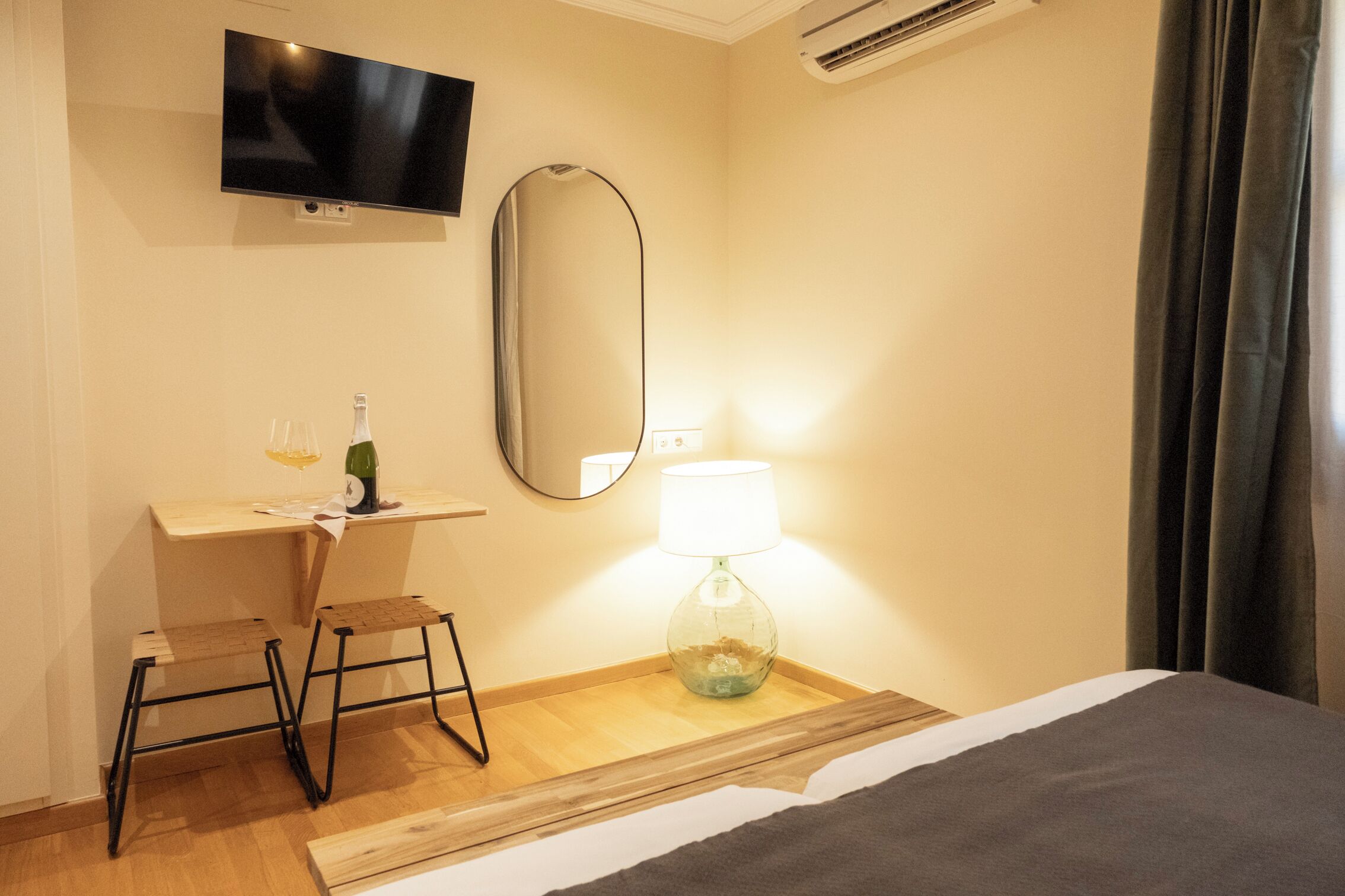 A hotel room with a bed, a desk with a chair, a television, a mirror, a lamp, and a bottle of wine.