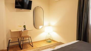 A hotel room with a bed, a desk with a chair, a television, a mirror, a lamp, and a bottle of wine.