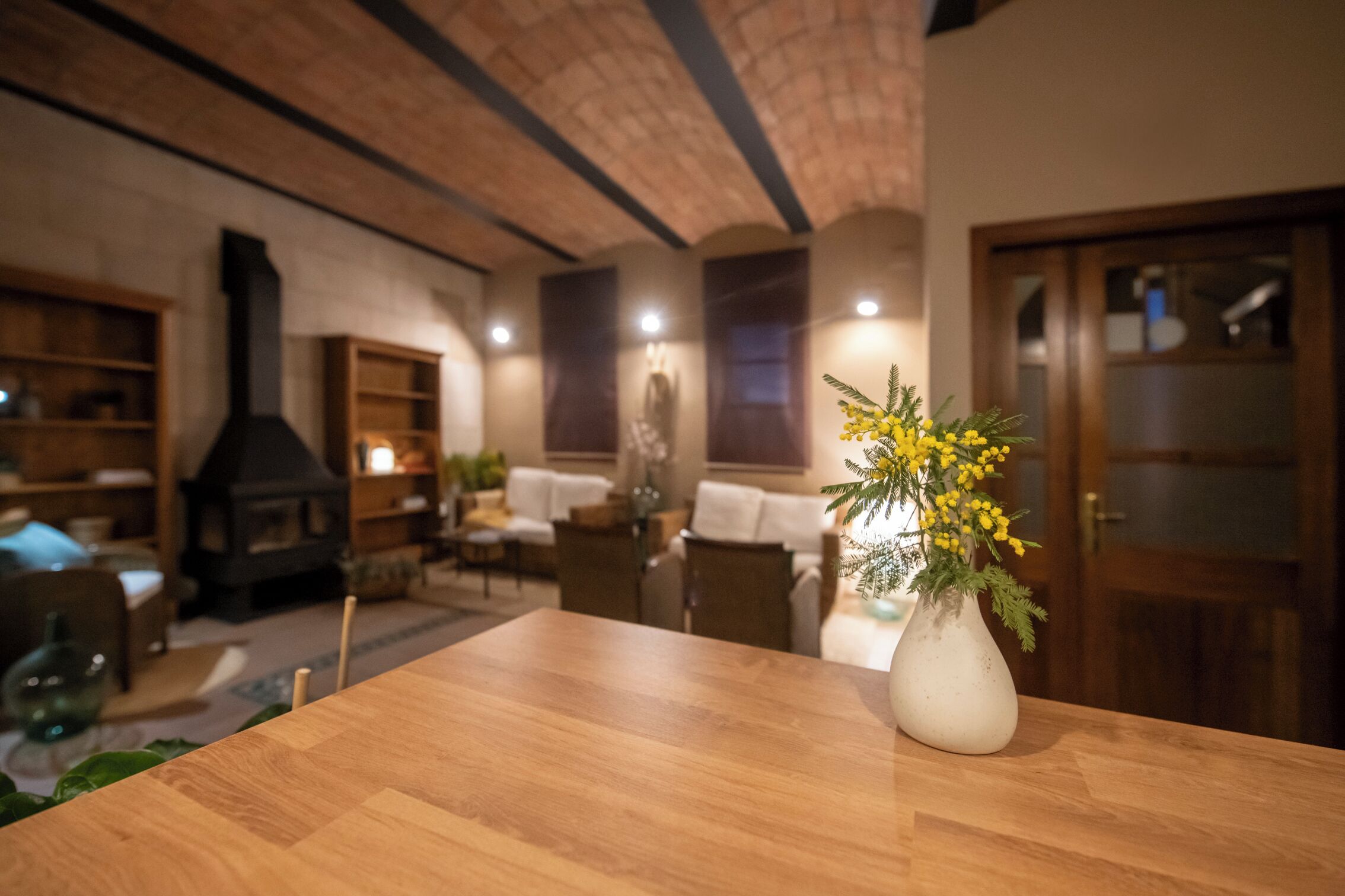 A wooden table with a white vase of yellow flowers in a living room with a fireplace, wooden shelves, and a door with frosted glass panels.