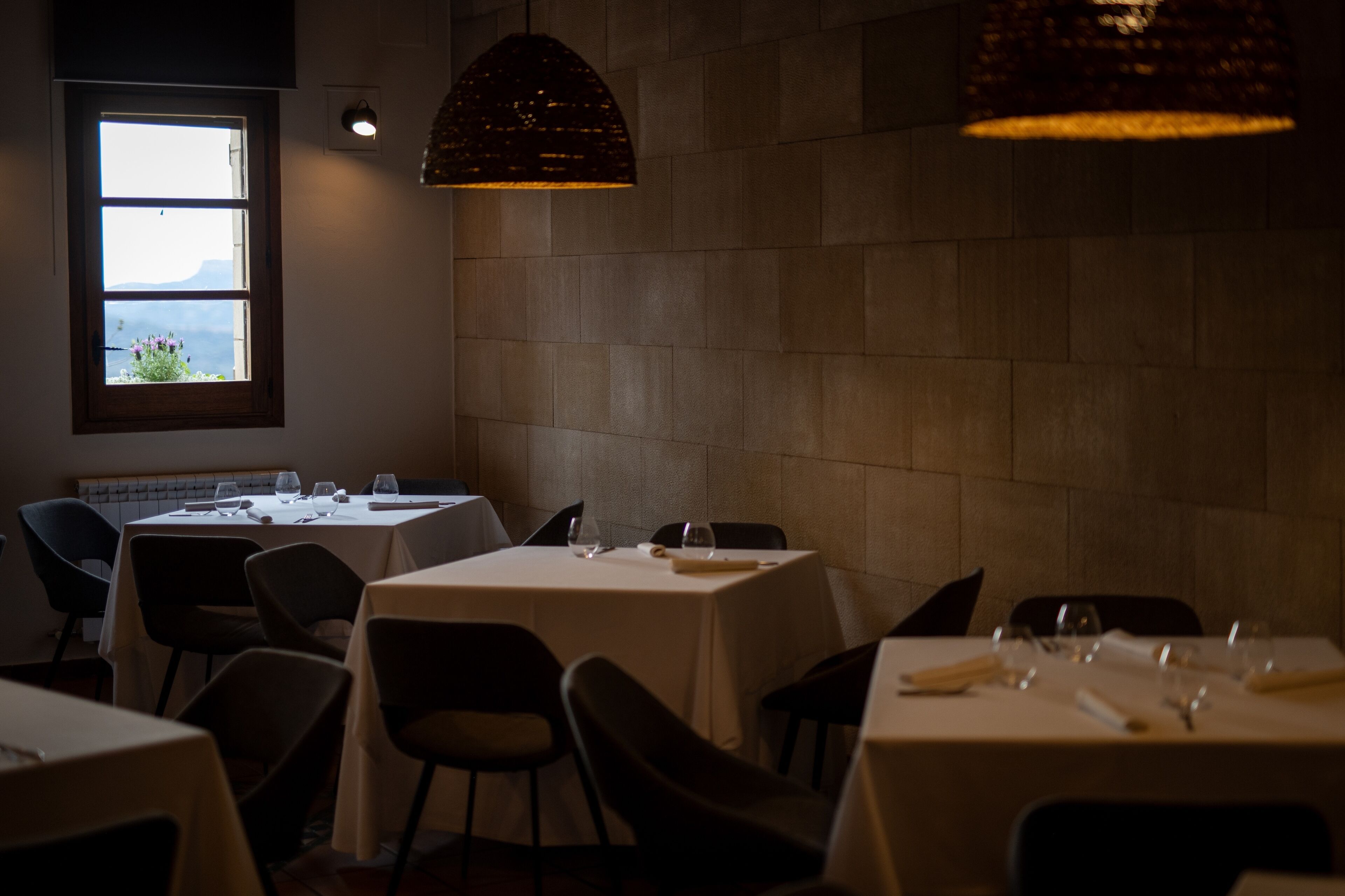 Ein schummrig beleuchtetes Restaurant mit gedeckten Tischen, einem Fenster mit Blick und einer strukturierten Wand.