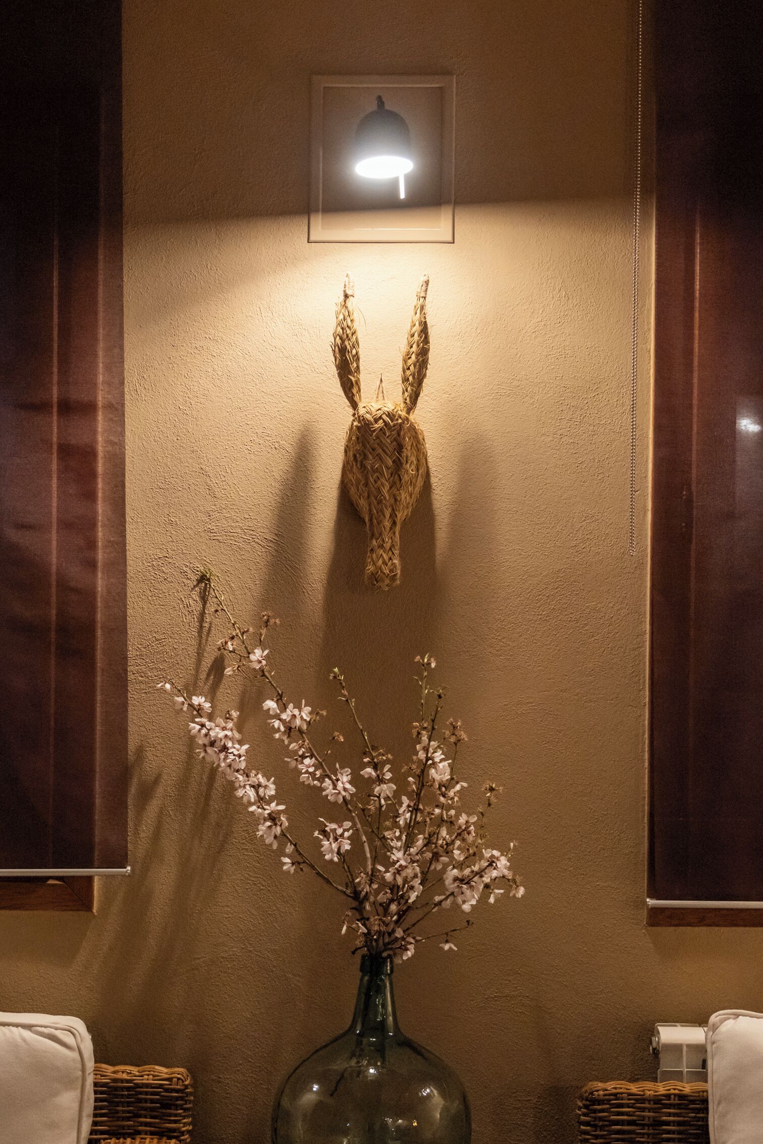 A bathroom with a woven animal head decoration, a vase with flowers, and a framed light fixture on the wall.