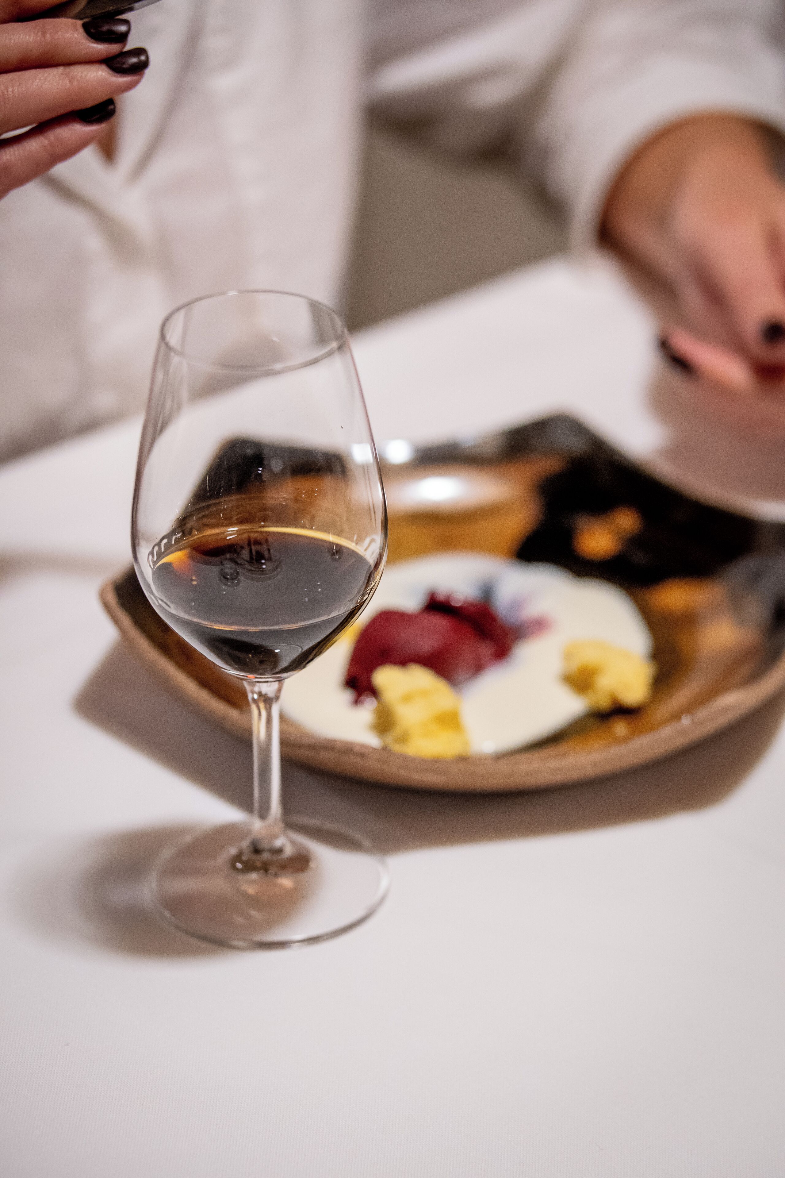 A close-up of a wine glass on a table with a plate of food in the background.