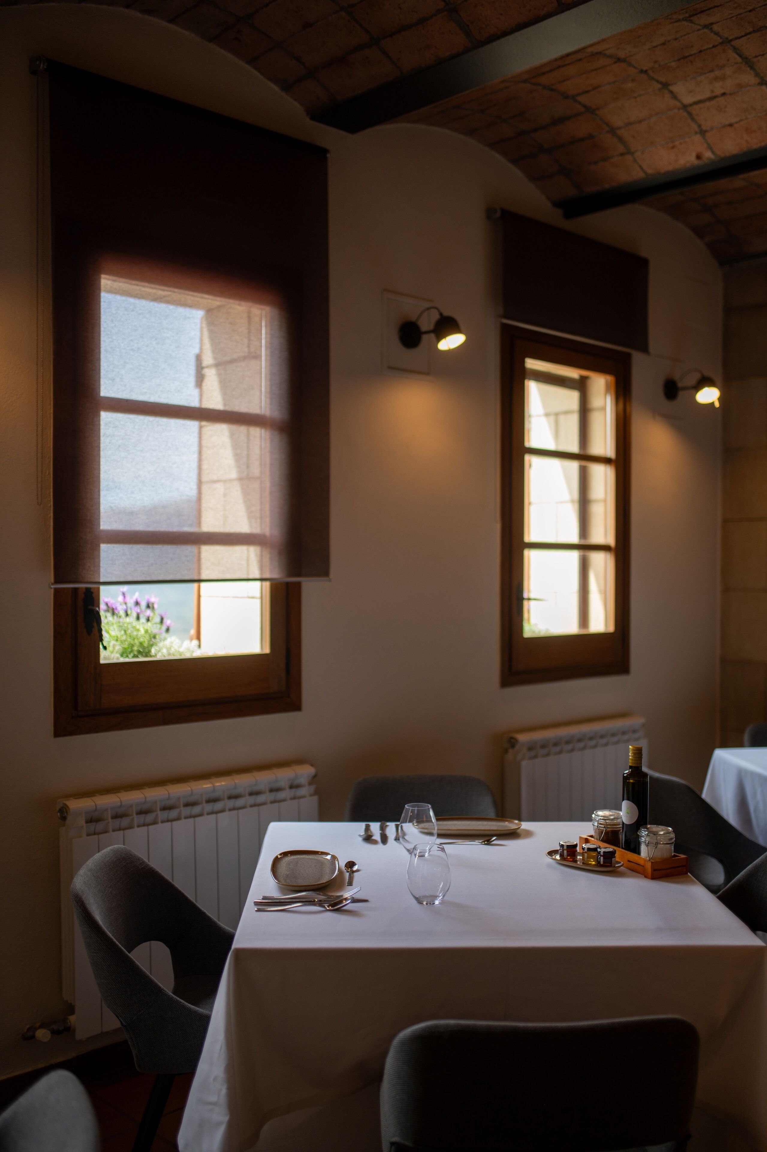 Een eetgedeelte met een tafel gedekt voor twee personen, twee ramen met zonwering, een radiator en een fles op de tafel.