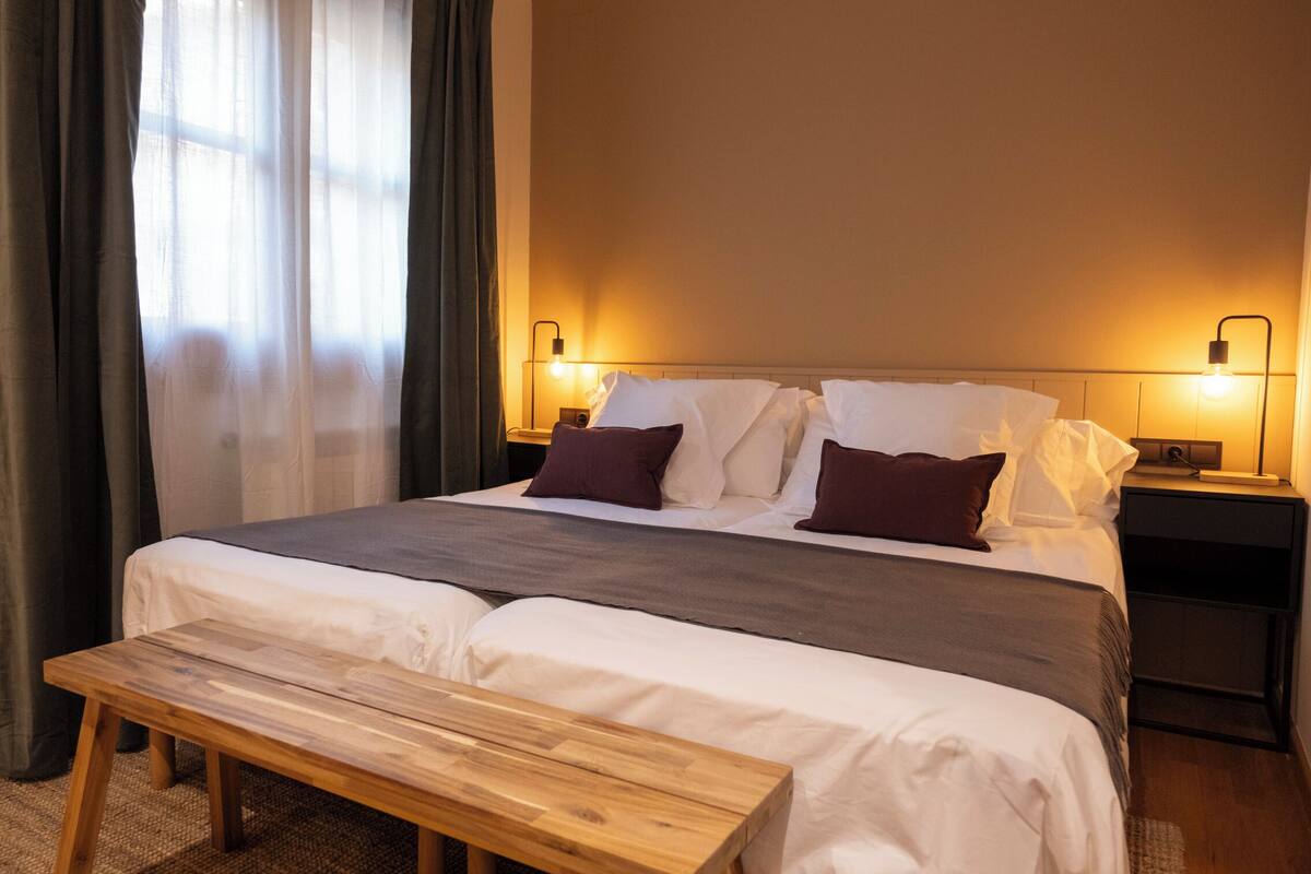 Ein ordentlich bezogenes Bett mit weißer und brauner Bettwäsche, eine Holzbank und zwei Nachttischlampen.
