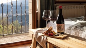 Una mesa de madera con una botella de vino, dos copas, un plato de queso y uvas, y una vista de montañas a través de la barandilla de un balcón.
