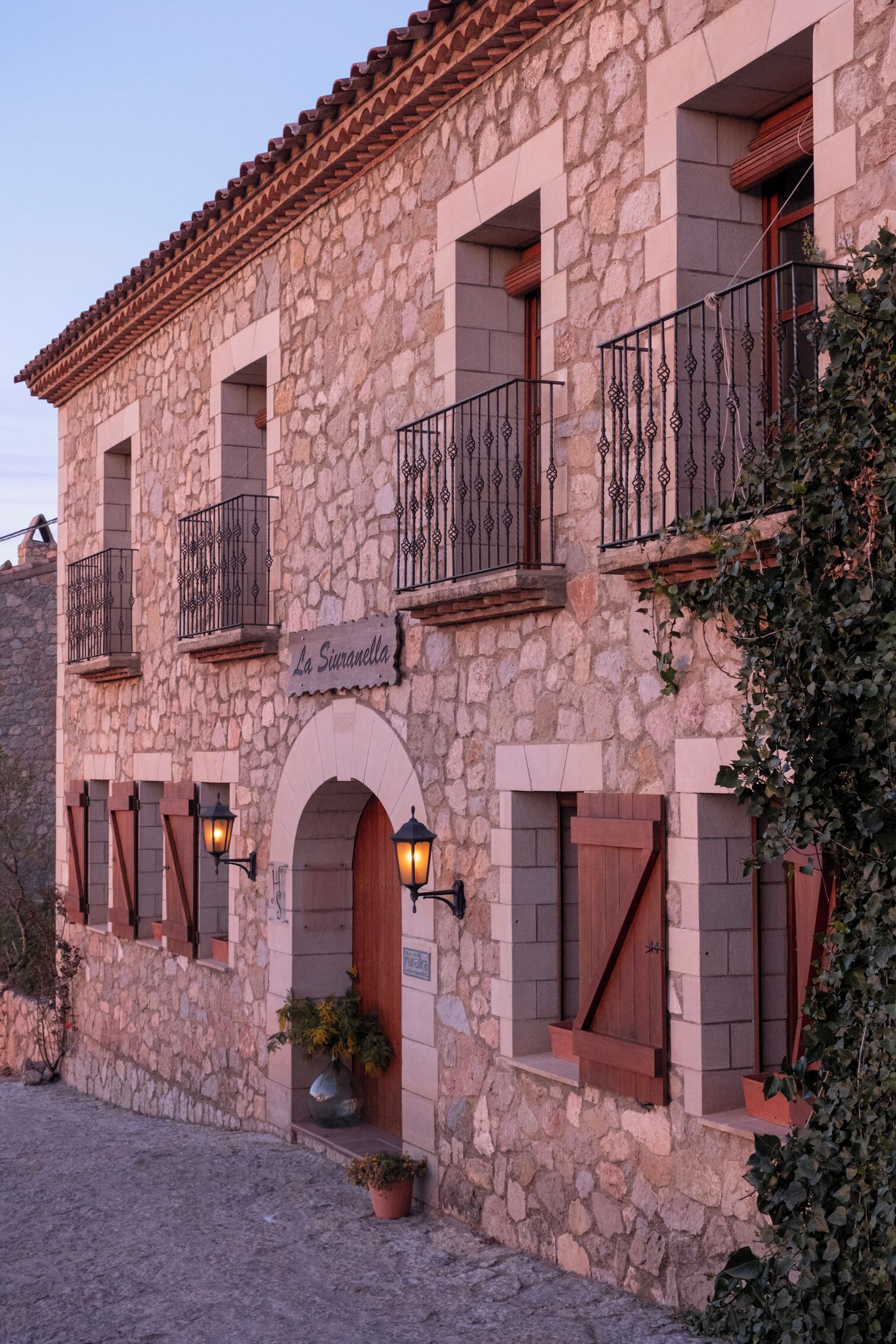 Un edificio de piedra con balcones, un letrero que dice "La Suranella" y plantas en macetas en la entrada.