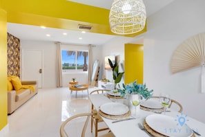 Un comedor luminoso con una mesa blanca dispuesta para cuatro personas, rodeado de paredes amarillas y una lámpara trenzada grande.