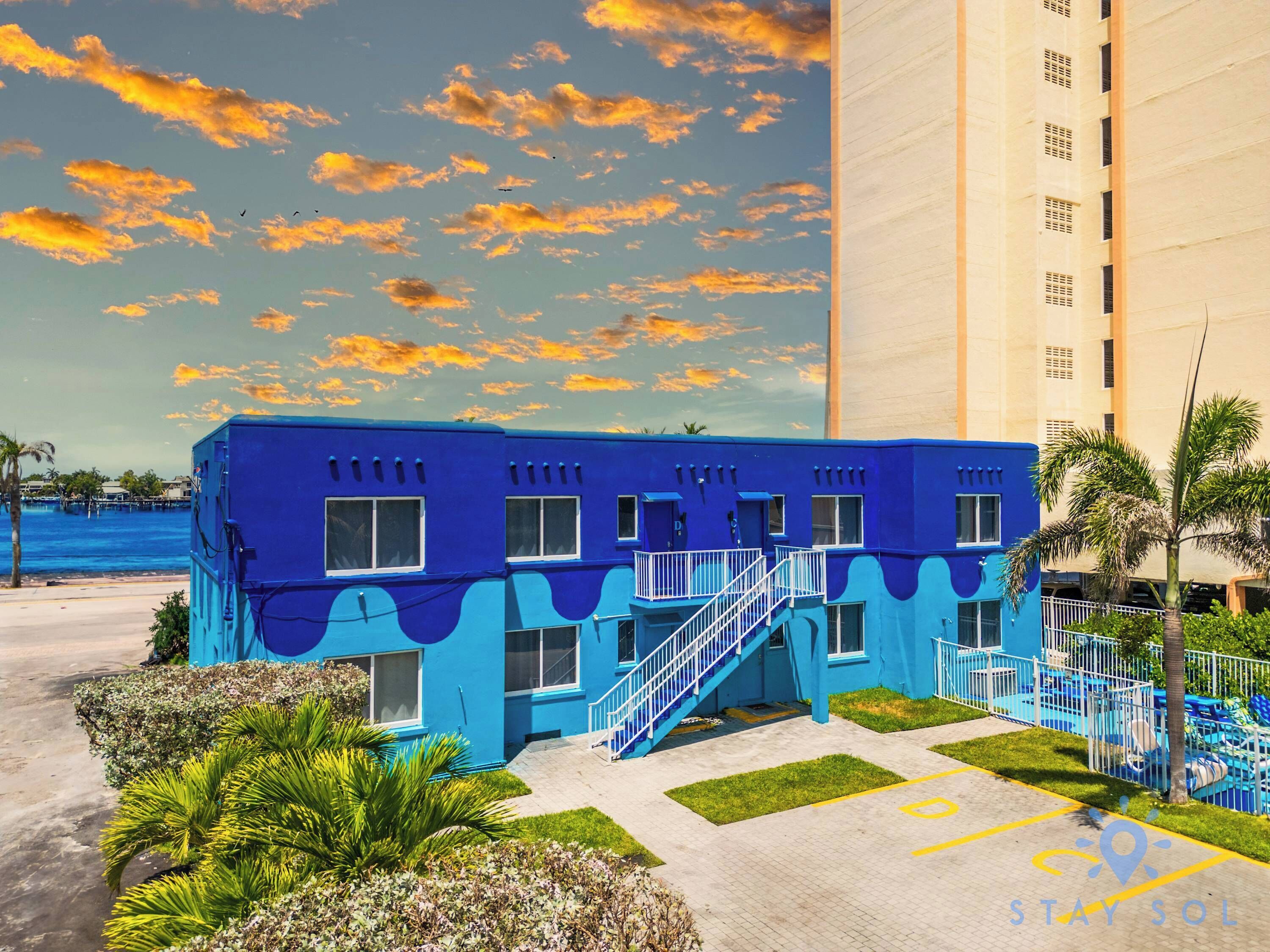 A blue building with multiple windows and a staircase leading to a balcony, situated next to a tall building and a body of water.