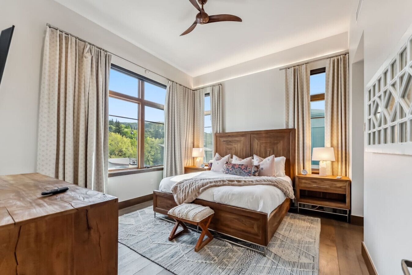 Une chambre avec un grand lit, un bureau en bois, un ventilateur de plafond et de grandes fenêtres avec des rideaux.