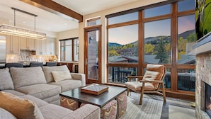 A spacious living room with a large window offering a mountain view, a cozy fireplace, and a modern kitchen in the background. - LUXE Riverfront Mountain Retreat | Walk to Gondola (Avon)