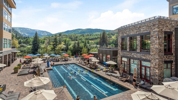 A pool area with lounge chairs and umbrellas, surrounded by a stone building with large windows, set against a mountainous backdrop. - LUXE Riverfront Mountain Retreat | Walk to Gondola (Avon)