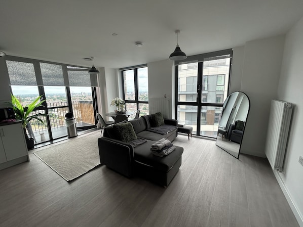 Un salon moderne avec un canapé noir, une table à manger avec des chaises, une grande fenêtre donnant sur des immeubles et un radiateur.