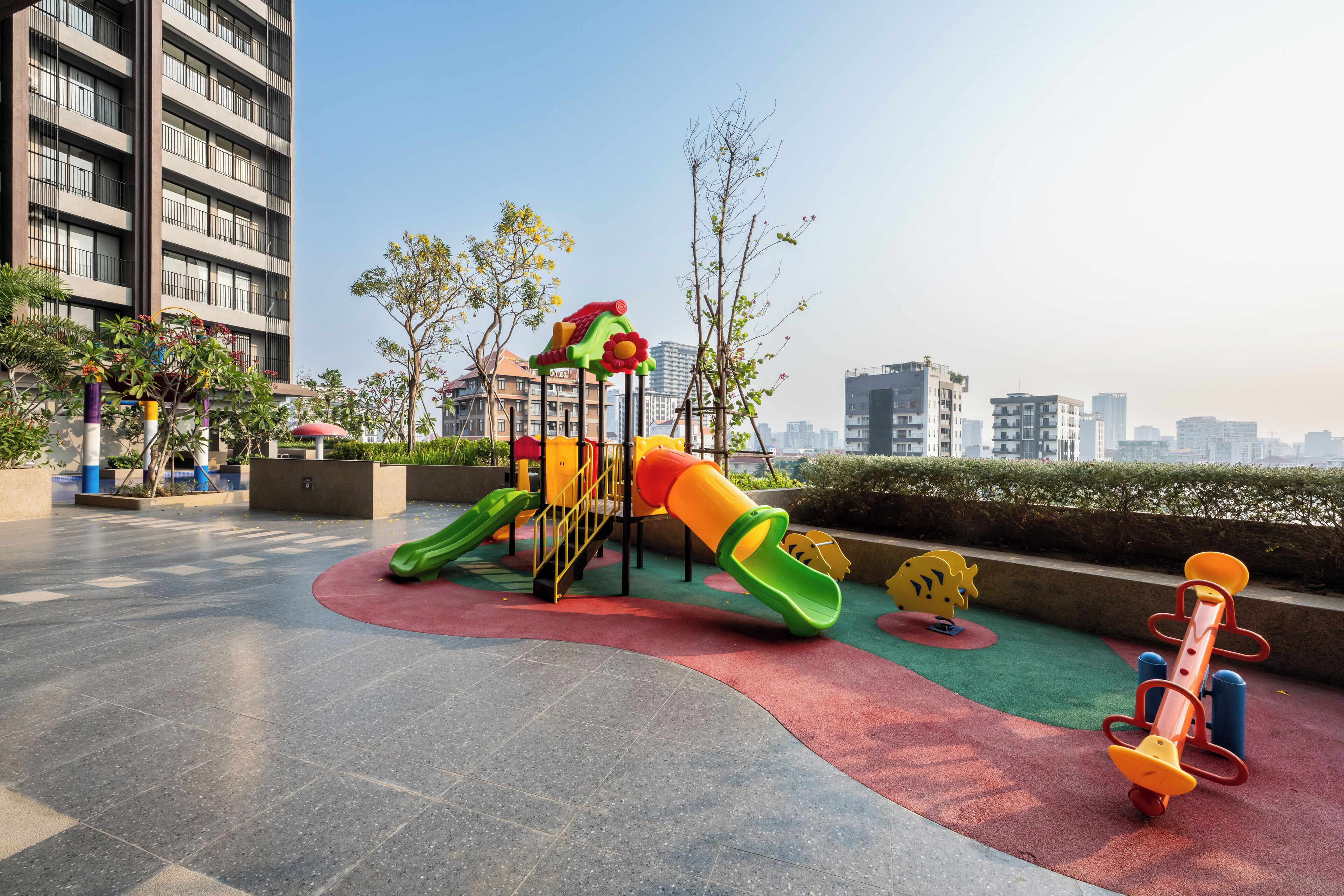Un parque infantil con toboganes y estructuras para escalar, rodeado de una zona pavimentada y edificios al fondo.
