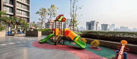 Un parque infantil con toboganes y estructuras para escalar, rodeado de una zona pavimentada y edificios al fondo.