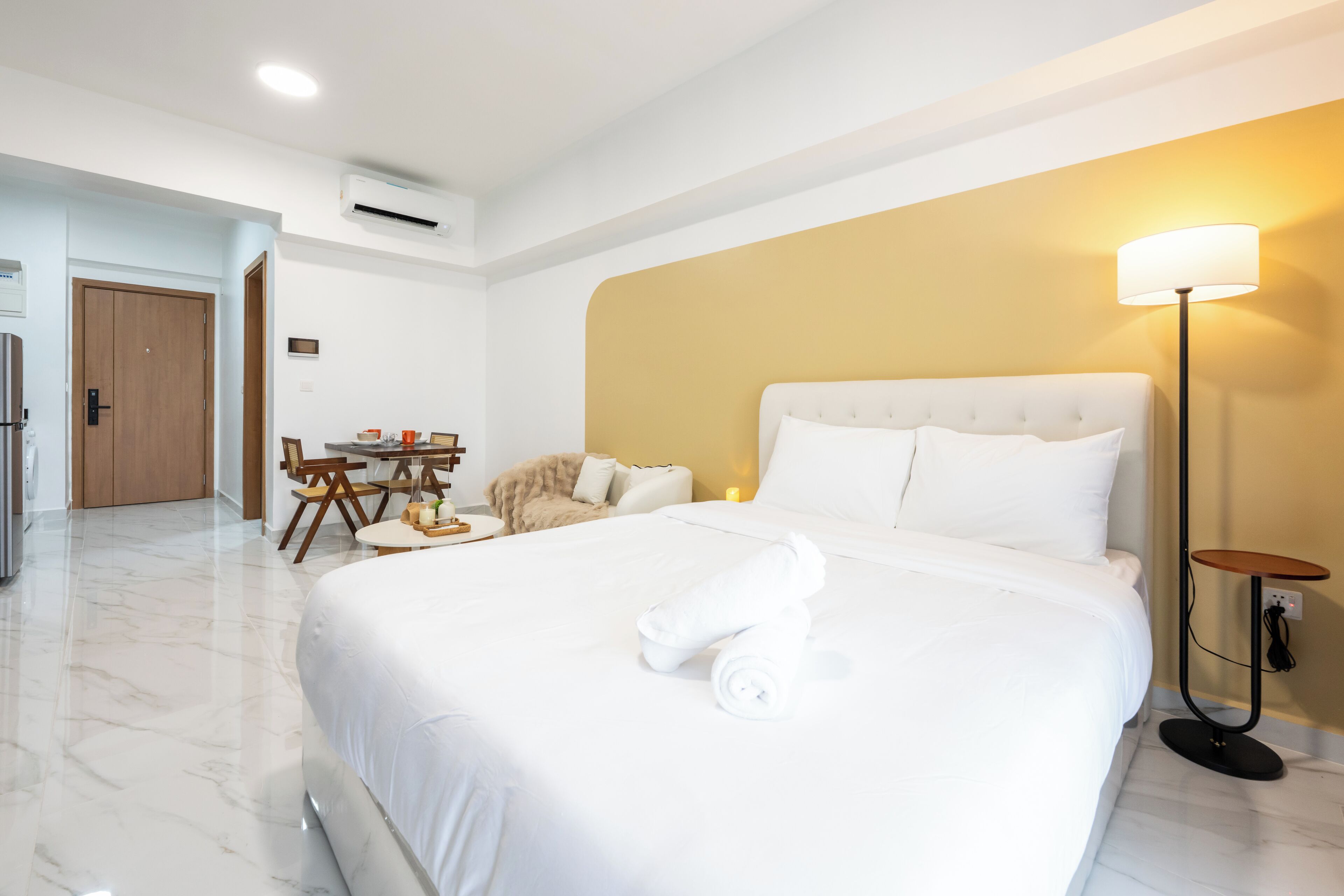 A modern hotel room with a large bed, a desk with chairs, a small table, and a standing lamp.