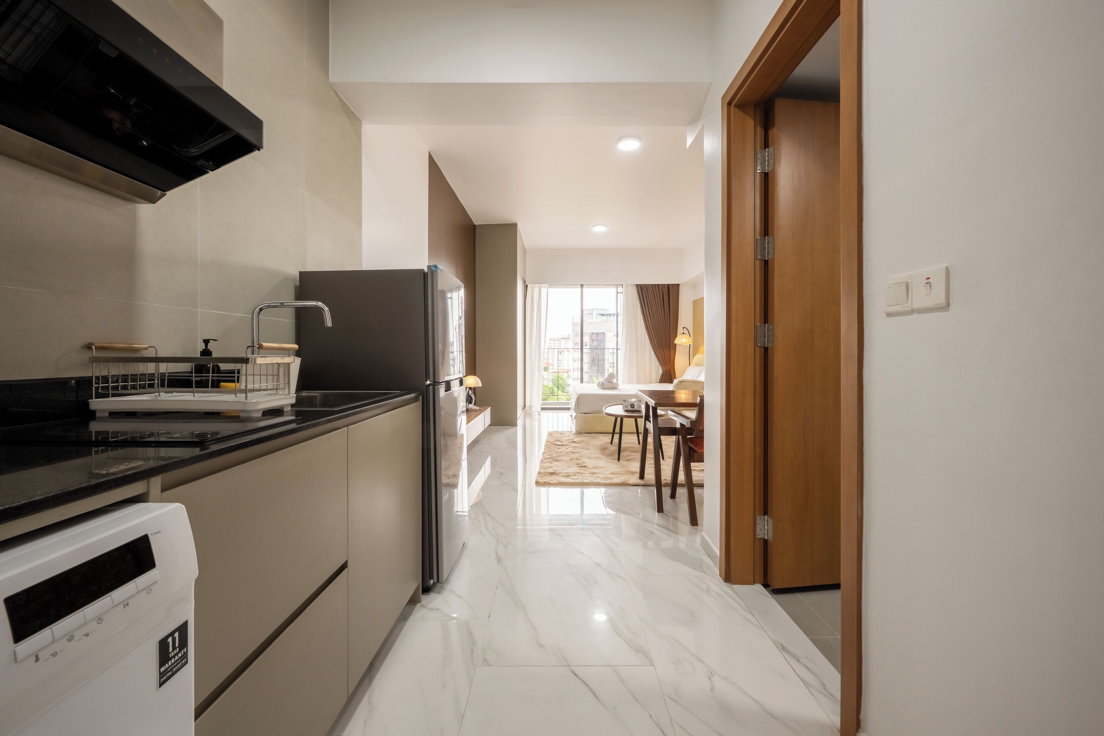 Una cocina moderna con encimera negra, electrodomésticos de acero inoxidable y suelo de mármol que conduce a una zona de comedor con mesa y sillas de madera.