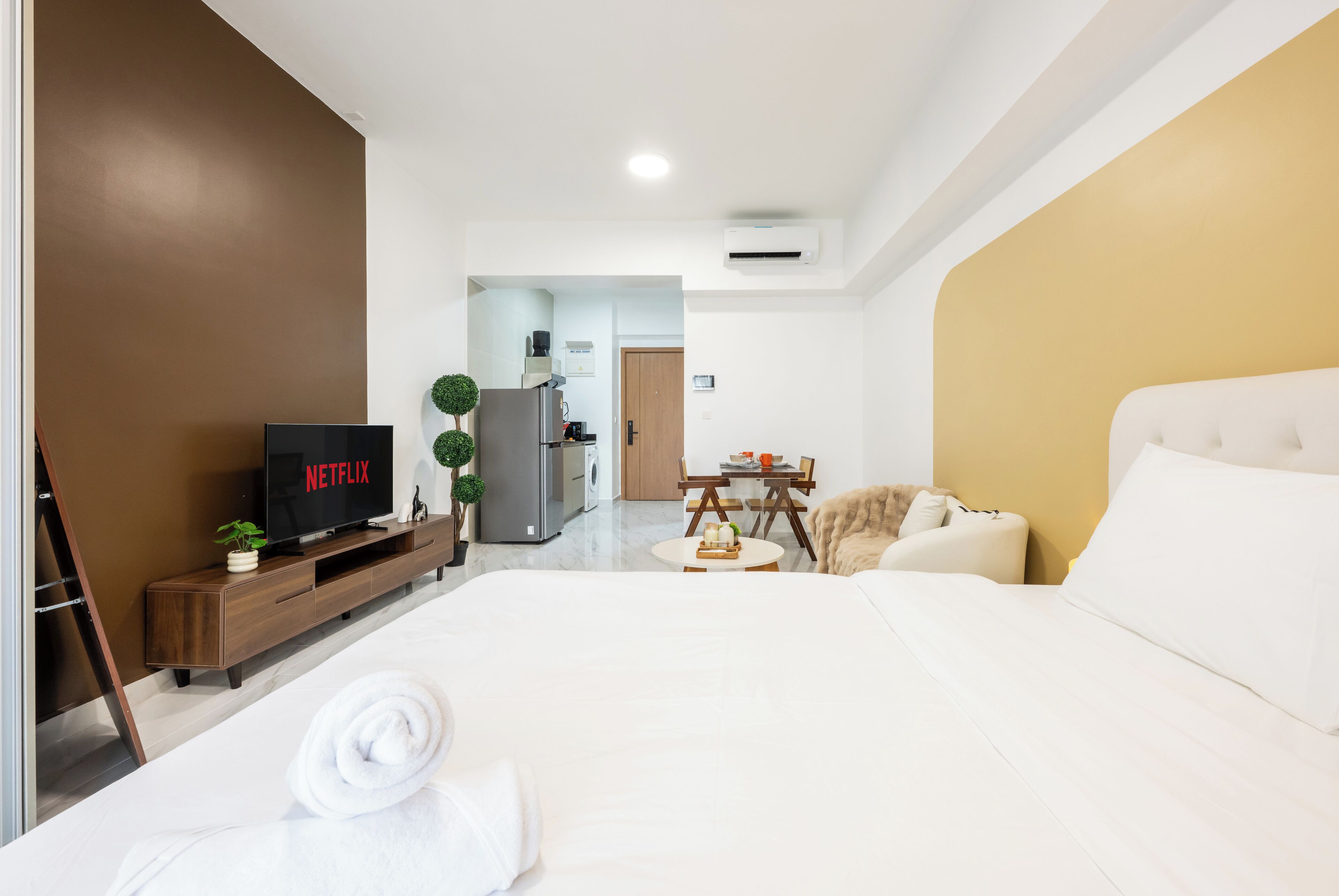 Habitación de hotel moderna con una cama grande, un televisor mostrando 'NETFLIX', una mesita con una planta y una kitchenette al fondo.
